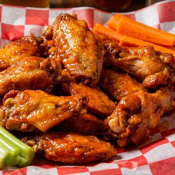 A red and white checkered basket filled with glistening buffalo wings, celery sticks, and carrot sticks.