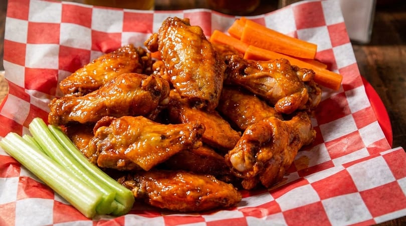 A red and white checkered basket filled with glistening buffalo wings, celery sticks, and carrot sticks.