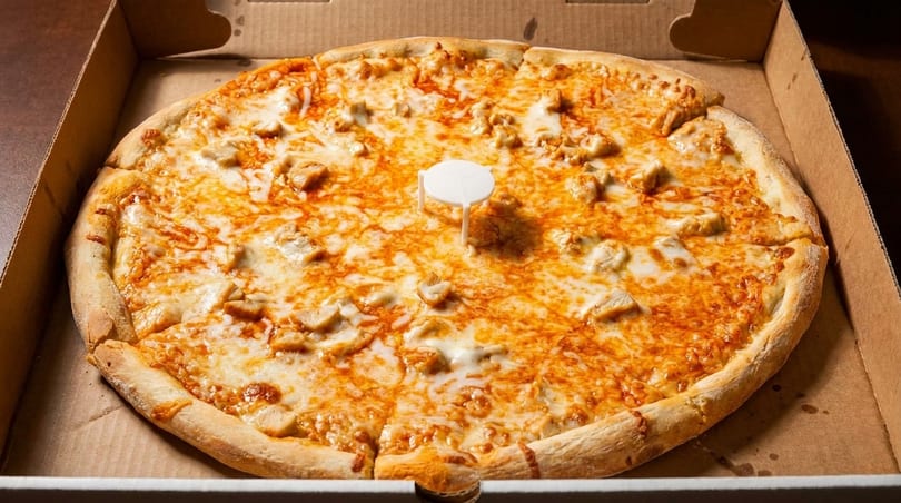 A whole chicken pizza with melted cheese and a pizza saver in a cardboard box.